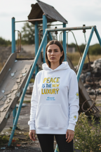 A woman stands in front of a ruined playground with rubble in the background, wearing a white crewneck sweatshirt with the text 'PEACE IS THE NEW LUXURY' in yellow and blue, and 'Allivood Zurich - Est. 2026' printed below.