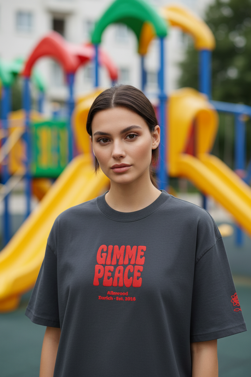 A woman stands in front of a colorful playground with yellow slides and blue structures in the background, wearing a dark charcoal gray oversized T-shirt with bold red text reading 'GIMME PEACE' on the front.