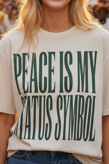 A smiling blonde woman stands in a city street during a parade or festival, wearing an oversized cream T-shirt with large dark green text reading 'PEACE IS MY STATUS SYMBOL' and blue jeans. Buildings, a crowd, balloons, and confetti are visible in the background.