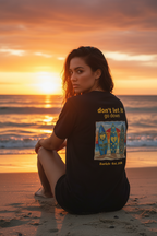 Woman sitting on a beach at sunset wearing a black t-shirt with a graphic design and text.