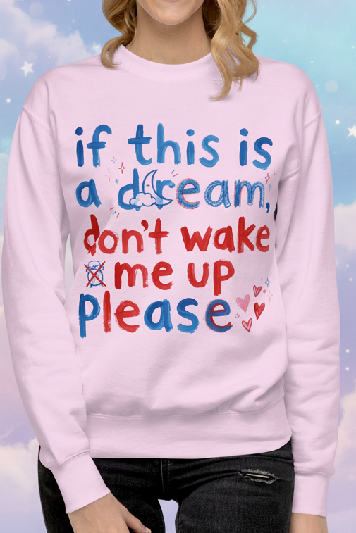A woman wearing a light pink crewneck sweatshirt with a colorful graphic text that reads 'if this is a dream, don't wake me up please' in blue and red, set against a dreamy pastel sky with clouds and stars background.