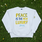 A white crewneck sweatshirt laid flat on green grass with small white flowers. The sweatshirt has a colorful graphic on the front reading 'PEACE IS THE NEW LUXURY' in yellow and blue lettering, with smaller blue text below.