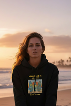 Woman sitting on a beach at sunset wearing a black hoodie with graphic design.