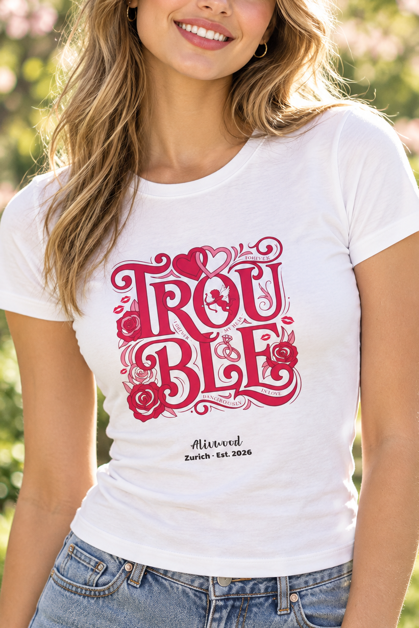 A smiling woman outdoors wearing a white short-sleeve T-shirt with a large pink decorative graphic reading 'TROUBLE' and smaller text below, paired with blue jeans. Trees with pink blossoms are blurred in the background.